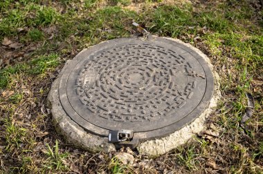 Çimlerin üzerinde çelik bir rögar kapağı. Sokakta gri betonun içinde bir tane siyah metal lağım kapağı var. Kanalizasyon kapağı küçük asma kilitlerle kapatılmış. Kanalizasyon kapağına iki kilit..