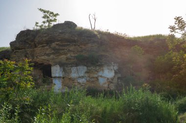 Kayalıklarda terk edilmiş mağara evi. Mağara eski şehir. Mağaralara giriş. Güneşin puslu ortamında inanılmaz bir manzara. Kayanın etrafındaki yemyeşil. Kayadaki mağaralara giriş..
