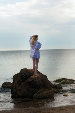 Kırmızı mayolu, beyaz gömlekli, tatlı bir sarışın, kayanın üstünde, arka planda duruyor. Genç kadın çıplak ayakla taş üstünde durduğunu hayal ediyor. Yaz ve tatil konsepti.