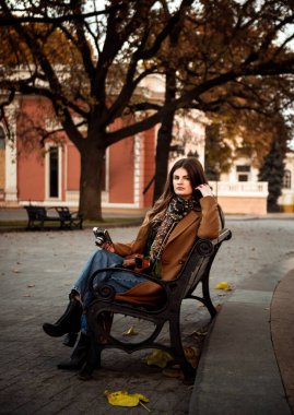 Kahverengi ceketli, kot pantolonlu ve siyah çizmeli manken sonbahar parkında bankta oturuyor. Eski mimarinin arka planına karşı. Elinde eski bir kamera, kameraya bakıyor.