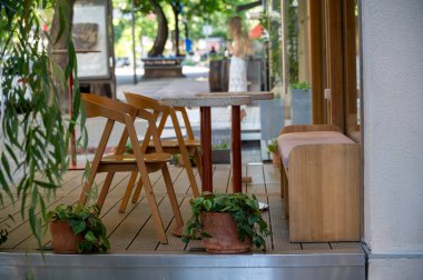Şehir merkezindeki yazlık terasta rahat bir kafeterya. Ön planda saksılar var. Güneşli bir günde kafede tahta sandalyeler. Arka planda bulanık insanlar.