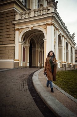 Uzun kahverengi ceketli, kot pantolonlu ve siyah çizmeli bir model mimari, dokulu bir kemerin arka planına karşı güvenle yürür. Elinde, sonbaharda yürüyen klasik bir kamera var..