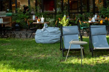 Çimlerin üzerinde güneş panelleri, yeşil park ağaçları ve dinlenecek bir yer olan bir yaz bahçesi. İki gri güneş şezlongu ve yazın dışarıda yuvarlak bir masa. Temiz havadaki çimenlerde dinlenmek için koltuk.