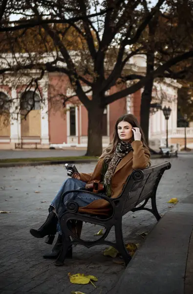 Kahverengi ceketli, kot pantolonlu ve siyah çizmeli manken sonbahar parkında bankta oturuyor. Eski mimarinin arka planında, fayansların üzerinde sarı sonbahar yaprakları. Elinde eski bir kamera, kameraya bakıyor..