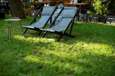 Çimlerin üzerinde güneş panelleri, yeşil park ağaçları ve dinlenecek bir yer olan bir yaz bahçesi. İki gri güneş şezlongu ve yazın dışarıda yuvarlak bir masa.