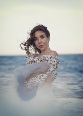 Elinde şifon kumaş tutan bir kadının portresi, rüzgarda savrulan saçlar, arka planda deniz ve gökyüzü, etrafındaki beyaz sis ve yansıma.