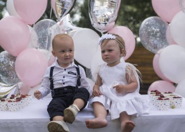 Küçük, şık giyimli çocuklar ilk doğum günlerini kutluyorlar. Erkek ve kız kardeş, ikizler balonların arka planında yan yana otururlar. Küçük prenses ve prens ilk doğum günlerinde