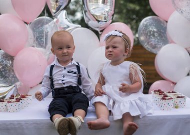 Küçük, şık giyimli çocuklar ilk doğum günlerini kutluyorlar. Erkek ve kız kardeş, ikizler balonların arka planında yan yana otururlar. Çocuklar açık havada parti veriyor.. 