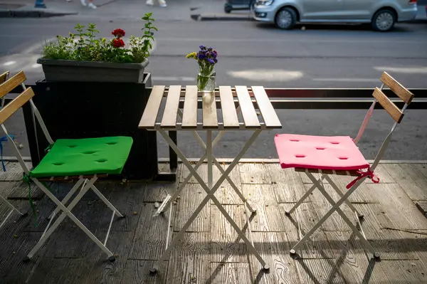 Sabahın köründe sokak kafesi şehrin arka planına karşı güneş ışınları altında. Boş teras, parlak sandalyeler ve masanın üzerinde cam şişeler içinde yabani çiçekler..