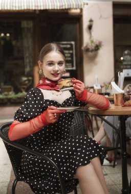 Parlak saç stili olan genç bir kadın bir kafede oturuyor. Gülümser ve altın bir fincandan kahve içer. Beyaz benekli ve kırmızı eldivenli siyah bir elbise giyiyor..