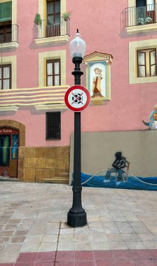 A Forbidden to play football sign stands in front of a vibrant pink building, with a mural depicting a religious figure and a black-and-white silhouette of a person on a boat. clipart