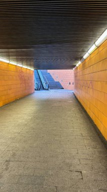 Parlak ışıklandırması olan sessiz bir yeraltı tüneli ve bir sonraki seviyeye çıkan bir yürüyen merdiven. Duvarlar turuncu fayanslarla süslenmiş ve atmosfer temiz ve modern hissettiriyor..