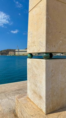 Stone structure with bronze spheres by the marina, contrasting with the blue sea and sky. A blend of modern design and coastal serenity with an industrial backdrop. clipart