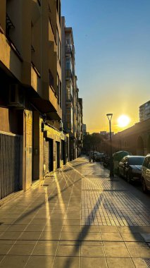 Batan güneşin altın ışıklarıyla aydınlatılmış sakin bir şehir caddesi. Uzun gölgeler kaldırım boyunca uzanıyor, sıcak ve huzurlu bir atmosfer yaratıyor..