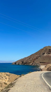 A serene coastal road winding along a rugged hillside, overlooking a tranquil bay with deep blue waters. The clear sky enhances the scene's peaceful charm. clipart