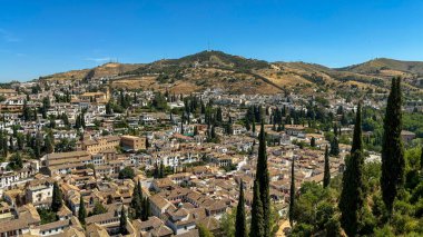 Granada 'daki Albayzn bölgesinin manzarası, açık mavi gökyüzü altında geleneksel beyazlatılmış evleri, selvi ağaçları ve yuvarlanan tepeleri sergiliyor..