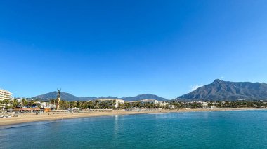 Marbella 'nın kumlu plajının güzel panoramik manzarası, arka planda ikonik La Concha dağı ve canlı mavi gökyüzünün altında sakin turkuaz sular..