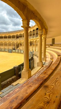Zarif taş kemerlerini, ahşap banklarını ve bulutlu bir gökyüzünün altında altın kum arenasını gösteren tarihi bir arenanın görüntüsü..