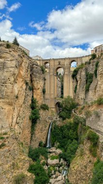 Derin bir kanyonu kaplayan çarpıcı tarihi bir taş köprü, yemyeşil bir yeşillik, çağlayan bir şelale, ve kabarık bulutlu parlak mavi bir gökyüzü. Ronda, İspanya.