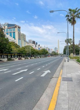 Gökdelenleri, palmiye ağaçları ve uzak mesafedeki ikonik Obelisk ile dolu kalabalık bir Buenos Aires caddesinin manzarası canlı şehir yaşamını ve modern mimariyi yansıtıyor..