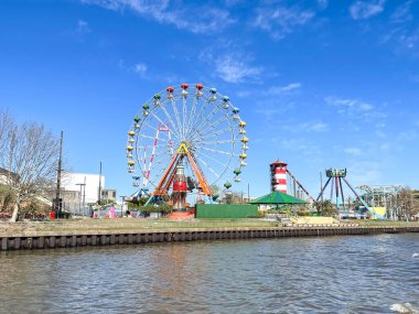 Renkli bir dönme dolap ve çeşitli eğlence araçları açık mavi bir gökyüzünün altında nehrin yanında duruyor. Canlı sahne, nehir kenarındaki eğlence parkındaki eğlenceli ve canlı bir atmosferi yansıtıyor..