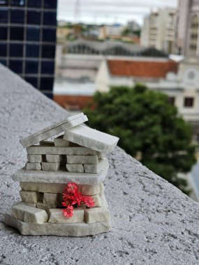 Miniature stone house with red flowers stands out against a blurred urban backdrop and greenery, blending creativity, nature, and urban elements under an overcast sky. clipart