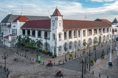 Kemerli pencereleri ve kırmızı çatısı olan tarihi sömürge binası, mavi gökyüzünün altında, kaldırım taşı sokak ve motosikletlerle çevrili..