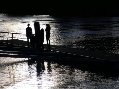 THE PONTOON OF L'ISLE-ADAM, digital horizontal colour photography: in the evening in France, le-de-France, Val d'Oise, a family of three people on a river pontoon with their silhouettes in the backlight, the father seeming to challenge something.