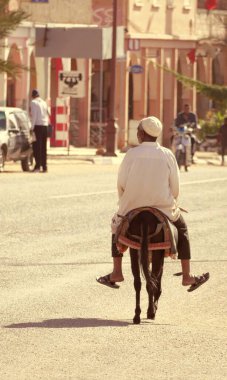 MAN ON DONKEY in MOROCCO, dikey dijital renkli fotoğraf: Zagora, Dra Valley, Dra-Tafilalet bölgesinde bir caddede tam gün ışığında, beyaz bir Chechia giyen bir adam eşeğinin sırtında, renkli yünüyle yol ortasında yürüyor