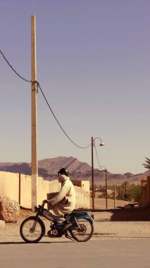 ZAGORA, FOCCO, MAGHREB, NORTH-AFRICA 'DA, küçük bir TRAVELLED yolda, dikey dijital fotoğraf: Parlak bir güneş ve mavi gökyüzü altında, bir adam bir djellaba ve bir kask giyen bir adam çevredeki bir duvar, dağlar ve elektrik hattı üzerinden mavi moped geçiyor.