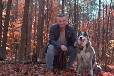 Orta yaşlı bir adam güz ormanında ağaç kütüğünde oturuyor. İri köpekli ve gülümseyen kameraya bakıyor.