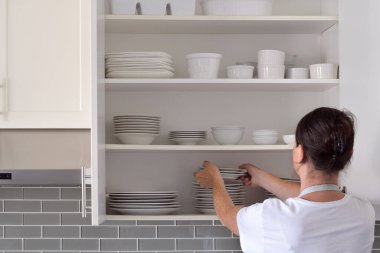 Kadın mutfak dolabından temiz beyaz tabakları yerleştiriyor ya da alıyor.