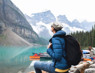 Sırt çantalı orta yaşlı kadın turkuaz gölü Moraine 'e renkli kanoları ve dağlarıyla bakıyor.
