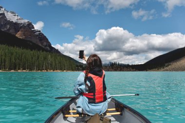 Genç kız, turkuaz mavisi Moraine gölünde, dağlarla çevrili bir kanoda kürek çekerken selfie çekiyor.