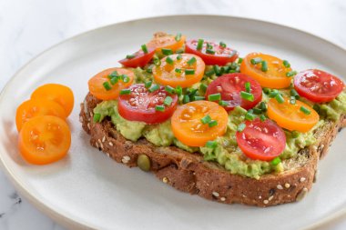 Yakın çekim çoklu tahıllı ekmek tostu, püre avokado ve renkli domatesler, sade taş tabakta soğanla süslenmiş.