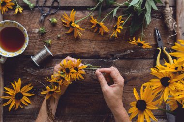 Kırsal ahşap masa üzerinde sarı çiçekli çelenk yapan kadınların el resimleri.