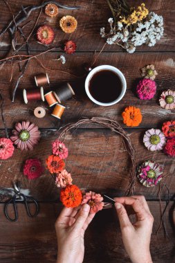 Kırsal ahşap bir masa üzerinde çiçek çelengi yapan kadın ellerinin yukarıdan çekimleri.