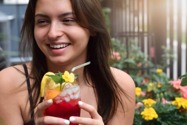 Mutlu genç kız gülümsüyor elinde renkli, kırmızı, soğuk bir yaz içeceğiyle bir bardak tutarken