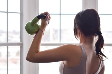 Evde pencerenin önünde çalışırken biseps olarak ağırlık kullanan genç bir kız.