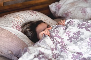 Genç kız yatakta uzanıp uyuyor, yüzünü battaniyeyle örterek rahatsız edilmek istemiyor.