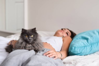 Yatakta gri kediyle yatan genç bir kadın kameraya bakıyor.