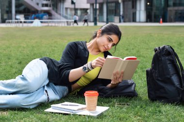 Yeşil çimlerde uzanan genç kız molada kitap okuyor.