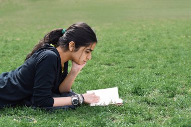 Yeşil çimenlerin üzerinde uzanmış kitap okuyan genç kız.
