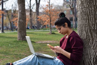 Parktaki ağacın altında dizüstü bilgisayarla oturan genç kız cep telefonu kullanıyor.