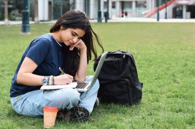 Genç kız çimlerin üzerinde dizüstü bilgisayar kullanarak oturuyor.