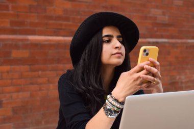 Siyah şapkalı genç kız kırmızı tuğlalı arka planda cep telefonu kullanıyor.