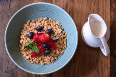 Mavi kase kahvaltılık granola taze çilek ve yaban mersini ve yanında yoğurt olan küçük beyaz seramik sürahi kırsal bir ahşap masa. 