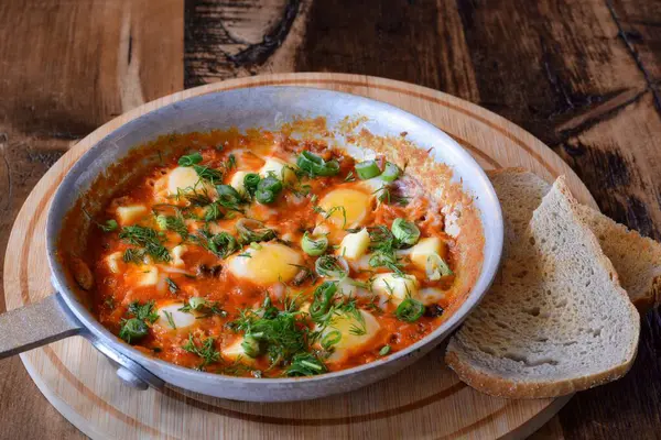 Taze dereotu ve yeşil soğanla süslenmiş kahvaltılık Shakshuka. Tahta masa üzerinde kızarmış ekmek ve kızarmış ekmek.