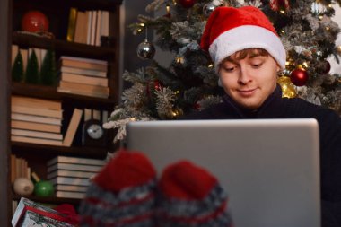 Noel Baba şapkalı genç adam dizüstü bilgisayar kullanarak Noel ağacında oturuyor.