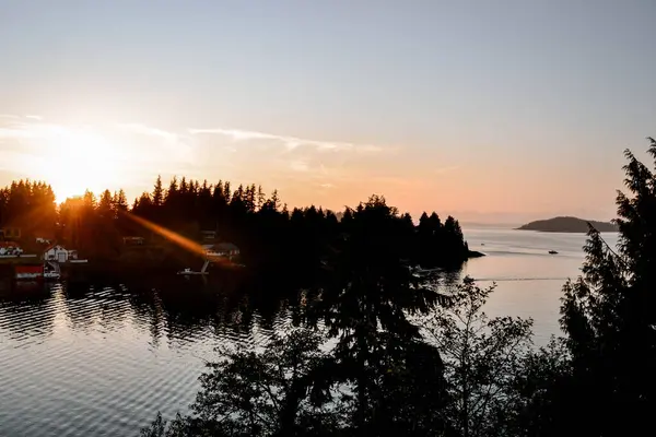 Altın bir günbatımı, sakin bir sahil kasabasının üzerine ılık renkler saçar. Sakin sular, ufku çevreleyen ışık ve sık orman siluetlerini yansıtır.. 